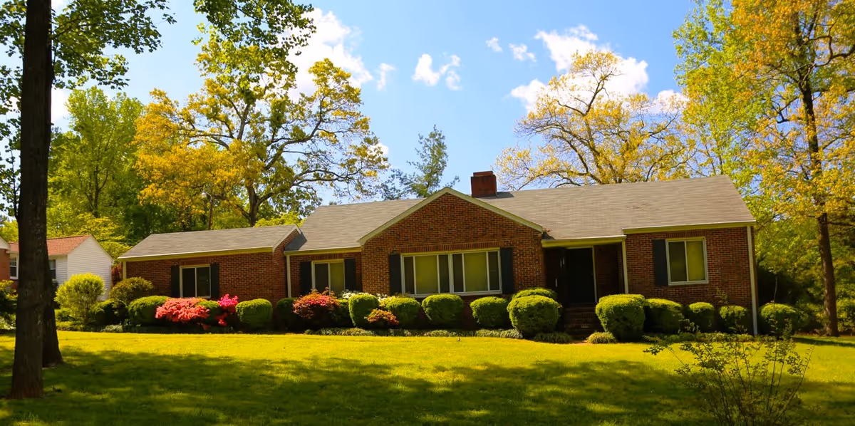 Single-story brick ranch house with manicured lawn, shrubs and large trees under a blue sky.