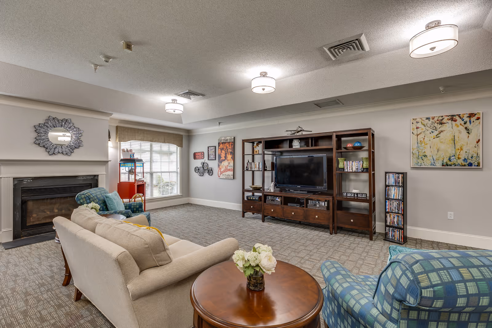 A cozy living room area with a beige sofa, two blue plaid armchairs, a round wooden coffee table with a flower vase, a fireplace with a decorative mirror above it, a large window with blinds, a red popcorn machine, a wooden entertainment center with a flat-screen TV, shelves with books and DVDs, and framed artwork on the walls.