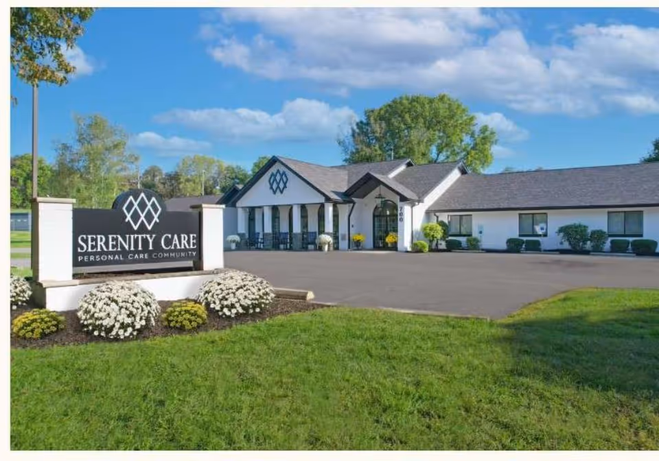 Exterior view of Serenity Care Kingston, a personal care community building with a large sign in front surrounded by landscaping and flowers, under a partly cloudy blue sky.