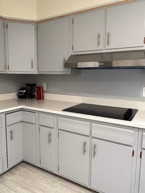 A clean kitchen corner with white countertops and light gray cabinets. There is a black electric stovetop with four burners and a stainless steel range hood above it. On the countertop, there is a black coffee maker and a red electric kettle. The floor has light-colored tiles.
