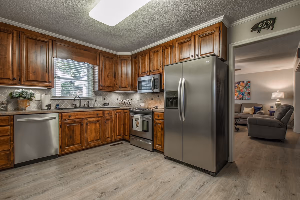 A kitchen with wooden cabinets, stainless steel appliances including a refrigerator, dishwasher, microwave, and stove. There is a window with blinds above the sink, and the floor is light wood. Through an open doorway, a living room with a sofa, armchair, lamp, and colorful wall art is visible.