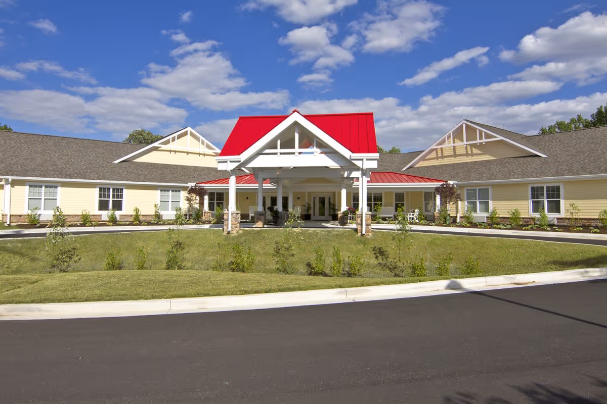 Front exterior view of a single-story senior living facility building with a bright red roof over the entrance, beige siding, and a well-maintained lawn under a partly cloudy blue sky.
