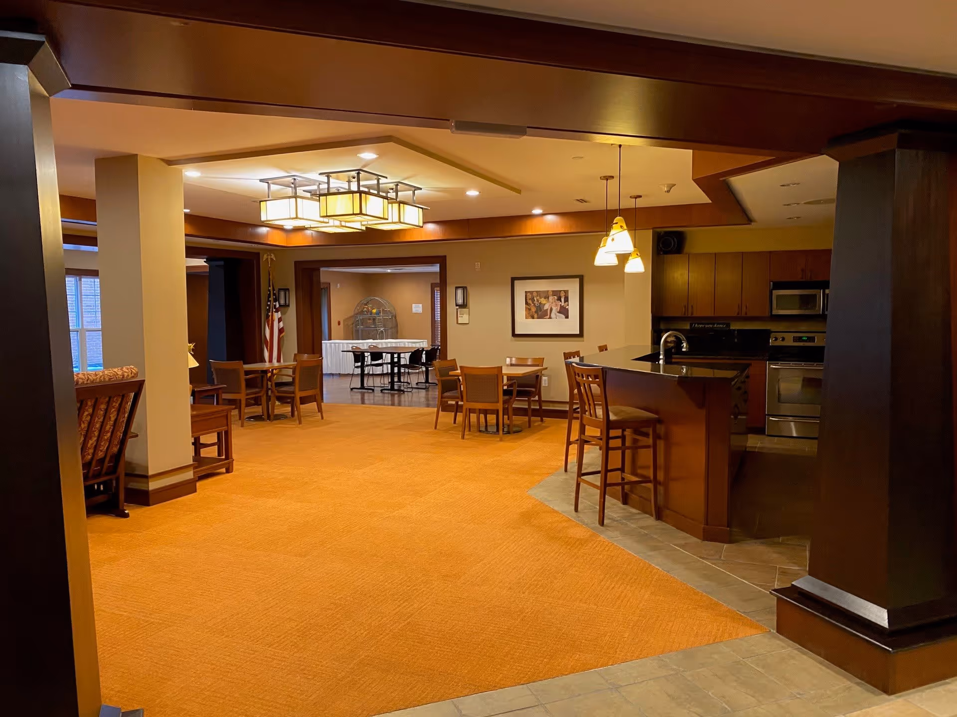 Interior view of a senior living facility common area with a kitchen and dining space. The room features warm lighting, wooden furniture including tables and chairs, a kitchen island with bar stools, and a carpeted floor. There is a framed picture on the wall and an American flag in the background.