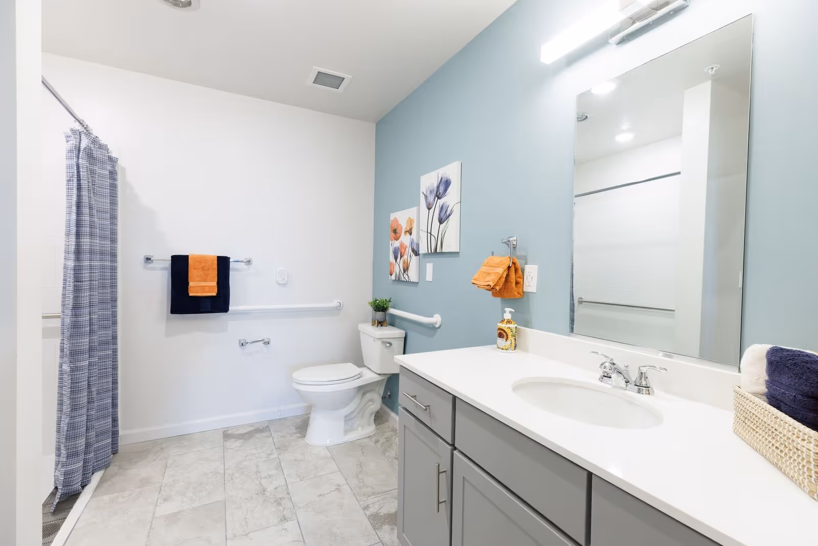 Bright and clean bathroom with a white toilet, a large mirror above a gray vanity with a white countertop and sink, a blue and white checkered shower curtain, and colorful towels hanging on the wall. The walls are painted white and light blue, with floral artwork above the toilet.