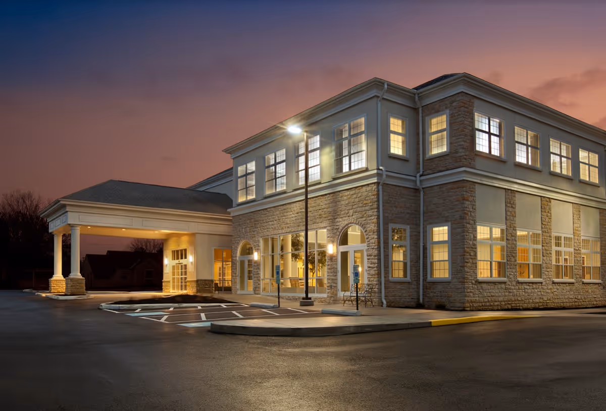 Exterior view of a two-story senior living facility building named Wesley Glen at dusk, with illuminated windows and a covered entrance area. The parking lot in front has designated handicap parking spaces and a street lamp is lit near the entrance.