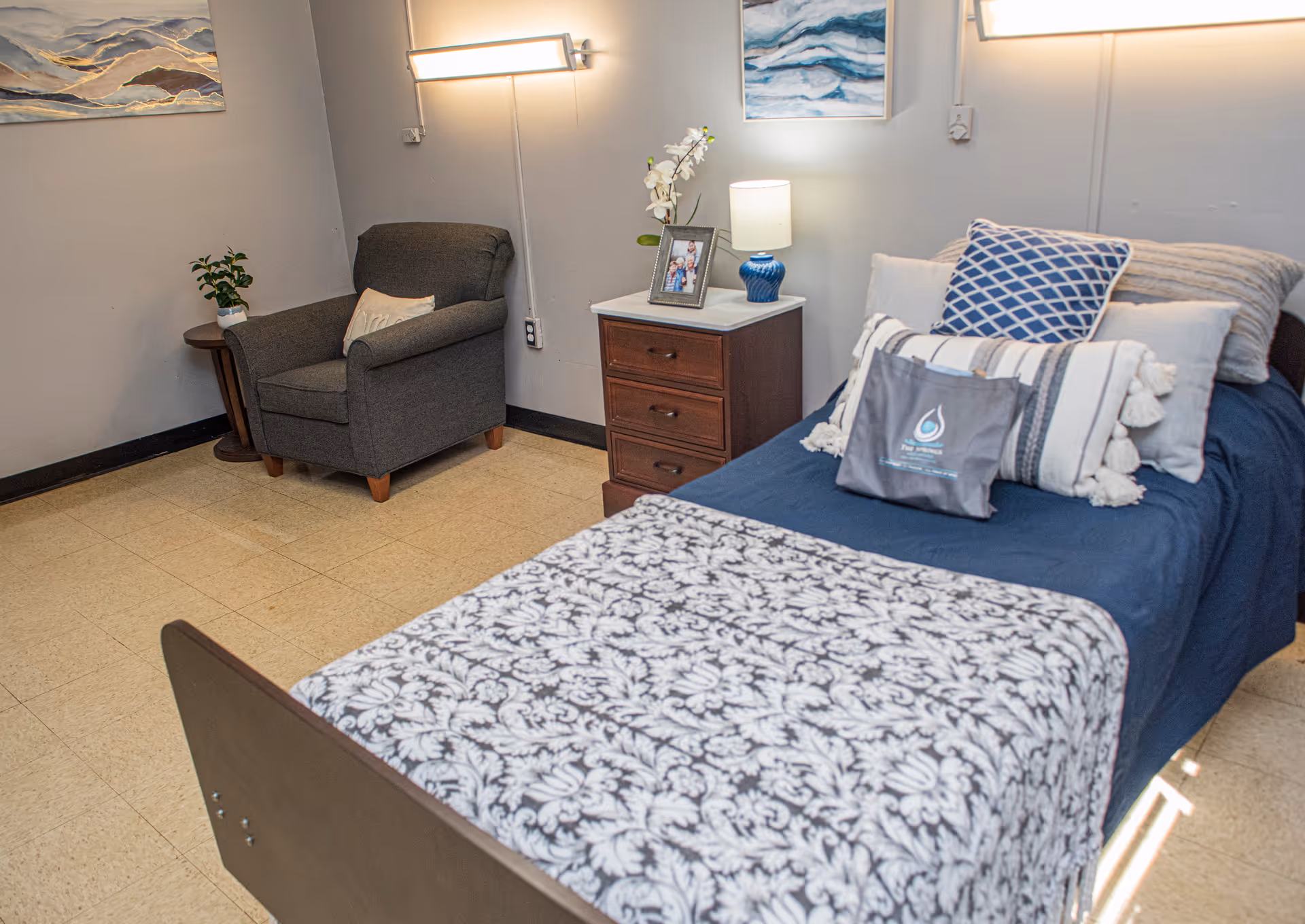 A neatly made single bed with multiple pillows and a patterned blanket in a room. Next to the bed is a wooden nightstand with a blue lamp, a framed photo, and a small plant on a round side table beside a gray armchair. The walls are decorated with two paintings and a wall-mounted light fixture.
