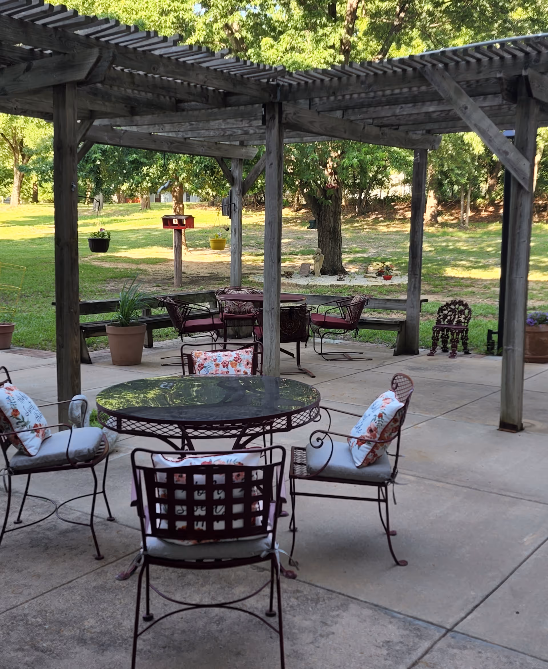Outdoor patio area with metal chairs and tables featuring floral cushions, under a wooden pergola. The patio overlooks a grassy area with trees and potted plants.