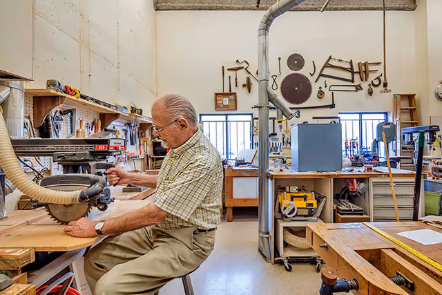 An elderly man works at a woodshop bench using a circular saw in a bright, tool-filled workshop.