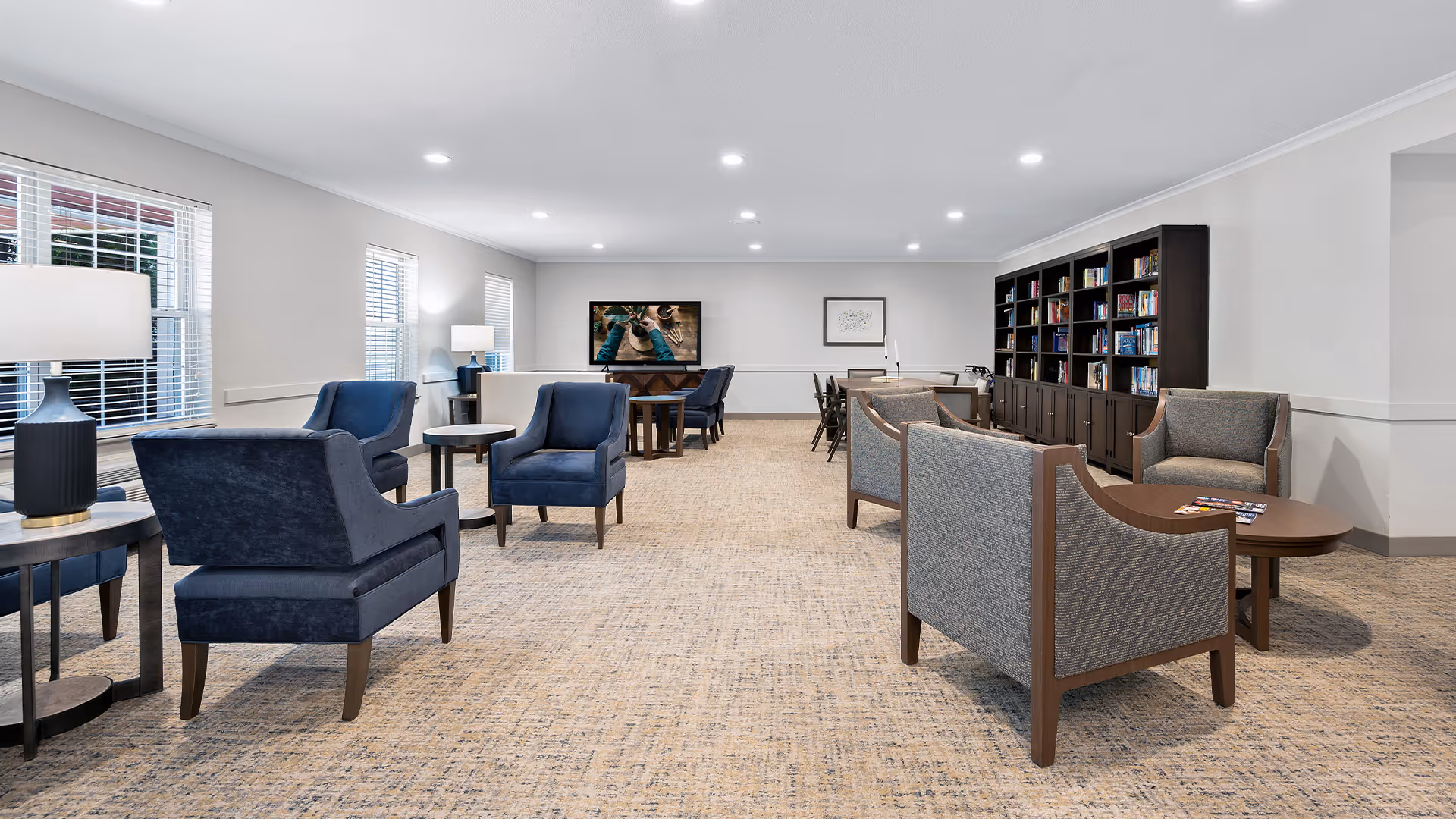 A spacious, well-lit common area with multiple seating arrangements including blue and gray armchairs and sofas around wooden tables. The room has large windows with white blinds, a carpeted floor, a bookshelf filled with books along one wall, and a television mounted on the far wall. There are also several lamps providing additional lighting.