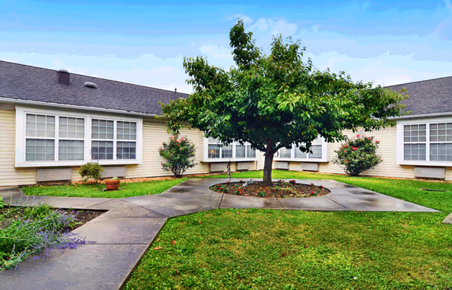 Outdoor courtyard area with a central tree surrounded by a circular flower bed and concrete walkways. The courtyard is enclosed by a single-story building with multiple large windows and beige siding. The sky is clear with some clouds.