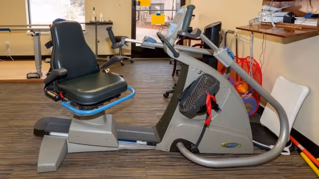 Indoor exercise room with a recumbent stationary bike in the foreground, a treadmill and other fitness equipment in the background, and a door leading outside. The room has wood-patterned flooring and light-colored walls.