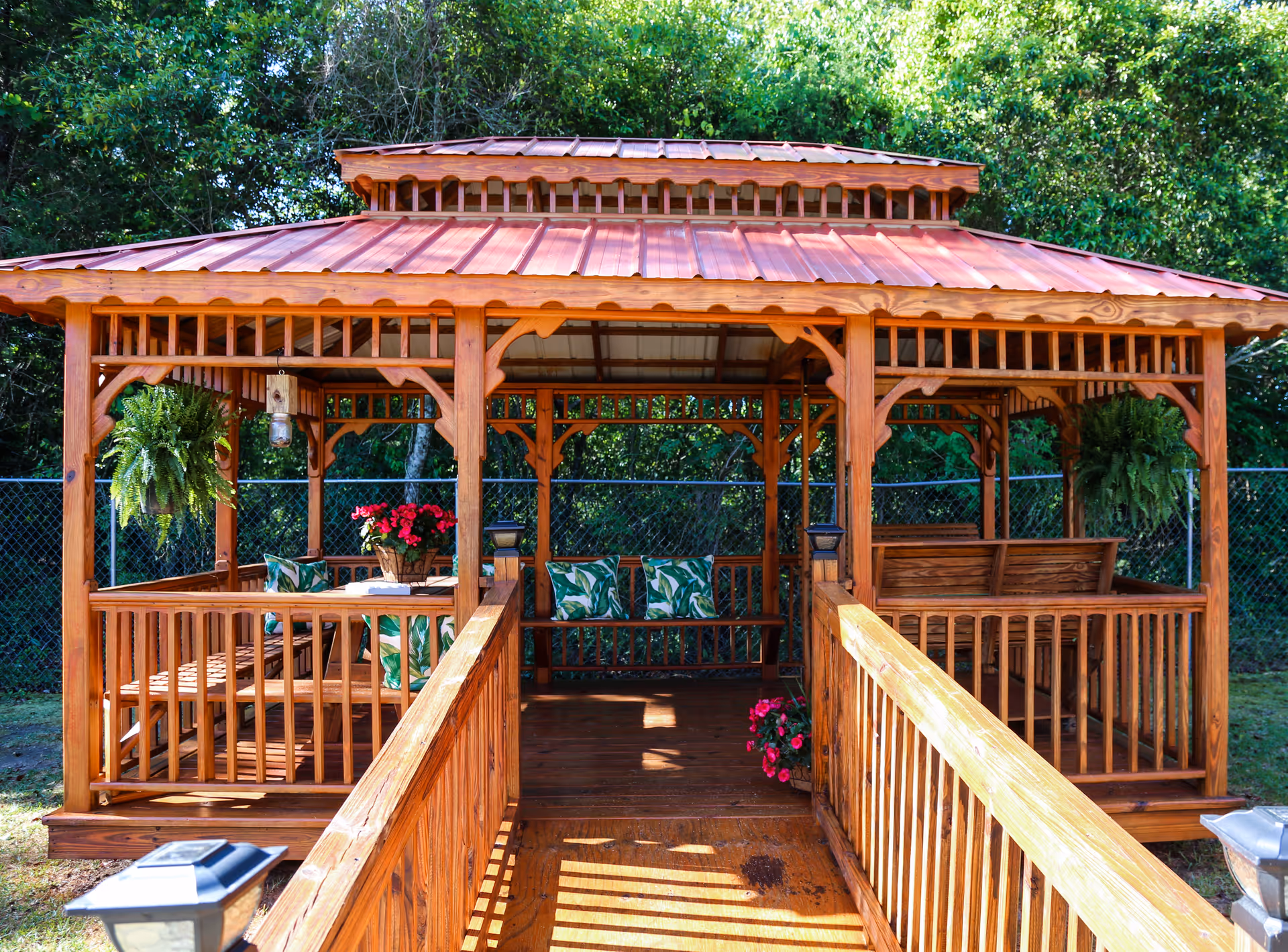 A wooden gazebo with a red metal roof situated outdoors, featuring cushioned benches with green leaf-patterned pillows, hanging ferns, and potted flowers. The gazebo is surrounded by trees and enclosed by a chain-link fence.