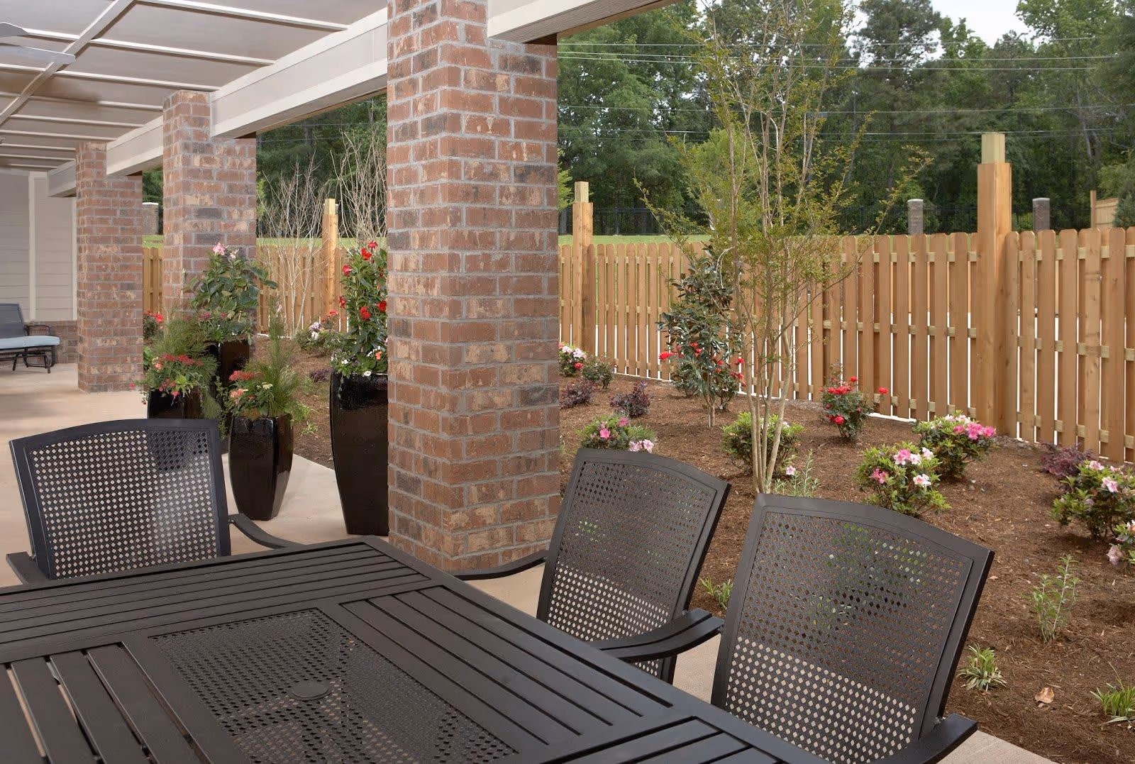 Covered outdoor patio area with black metal table and chairs, brick columns, potted plants with flowers, and a wooden fence enclosing a garden with small flowering bushes and trees.
