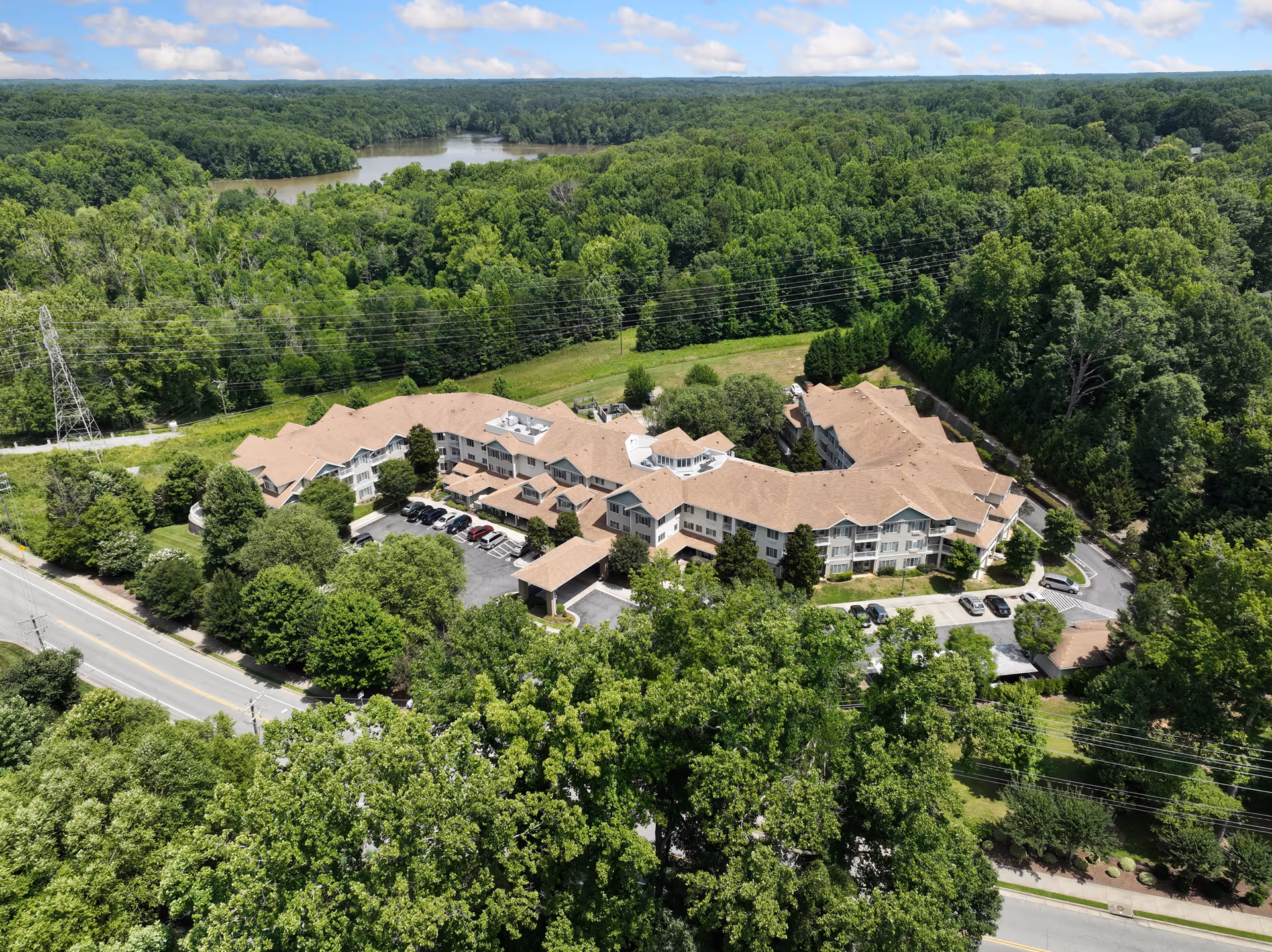Aerial view of Carolina Estates Gracious Retirement Living, a large multi-wing building surrounded by dense green trees and vegetation. There is a parking area with several cars in front of the building, and a river is visible in the background under a partly cloudy sky.