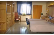 A rehabilitation patient room with a single bed, wooden storage cabinets, a chair by a window with blue curtains, and flowers on a table.