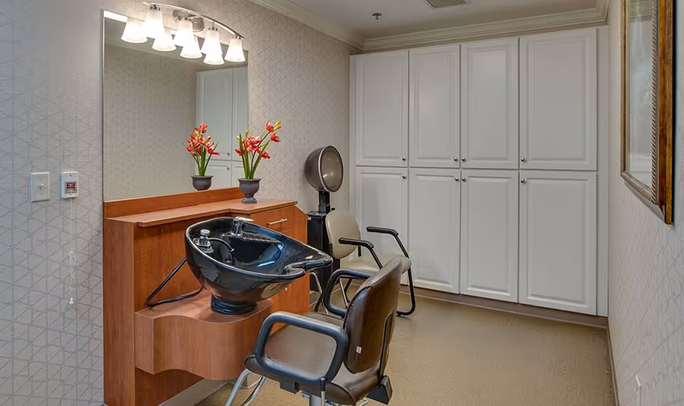 Interior view of a senior living facility hair salon area with a black hair washing sink, two salon chairs, a vintage hair dryer, a large mirror, a wooden cabinet with red flowers in vases, and white storage cabinets along the wall.