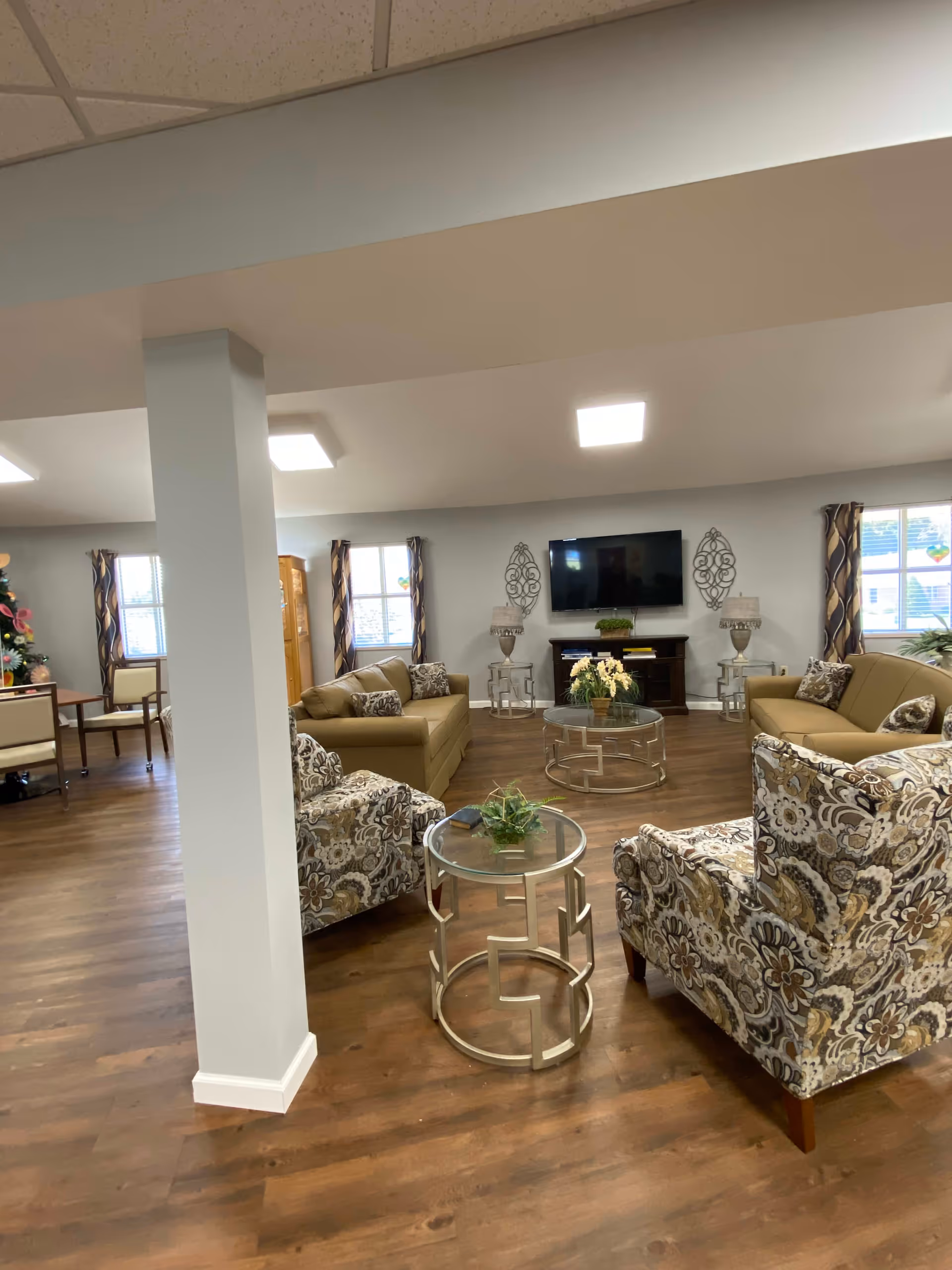 A cozy living room area in an assisted living facility with two patterned armchairs, two beige sofas, glass-top side and coffee tables with plants, a flat-screen TV mounted on the wall, decorative wall art, and windows with patterned curtains letting in natural light.