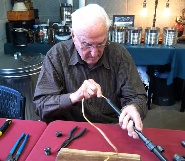 An elderly man with white hair and glasses is seated at a table covered with a red cloth, working with tools and a metal pipe. Various tools including pliers and a wrench are on the table. Behind him, there are several metal cans and other items on a table covered with a blue cloth.