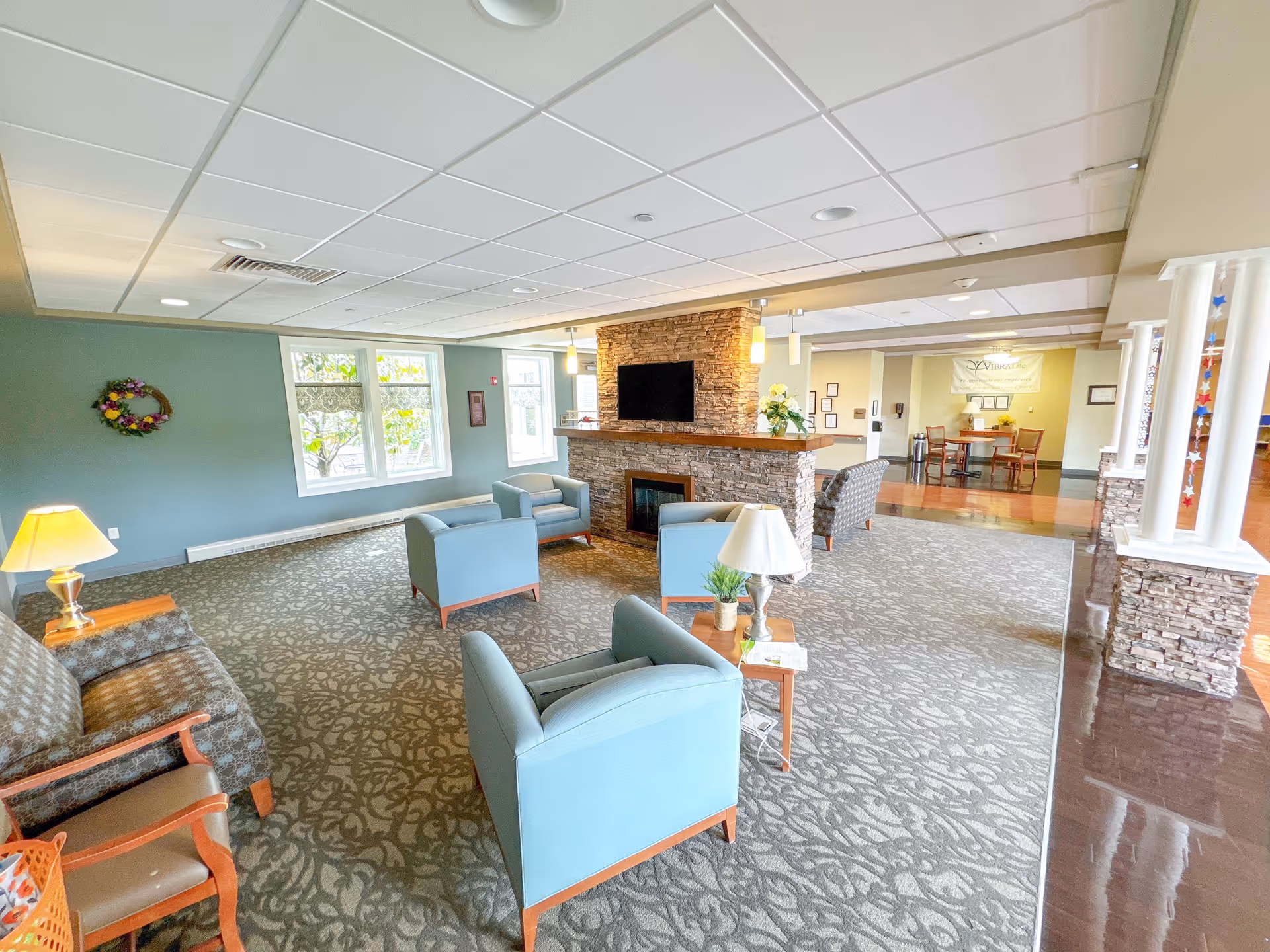 A spacious lobby area featuring comfortable seating, a stone fireplace, and large windows allowing natural light. Decorative elements include a wreath on the wall and a lamp on a side table.