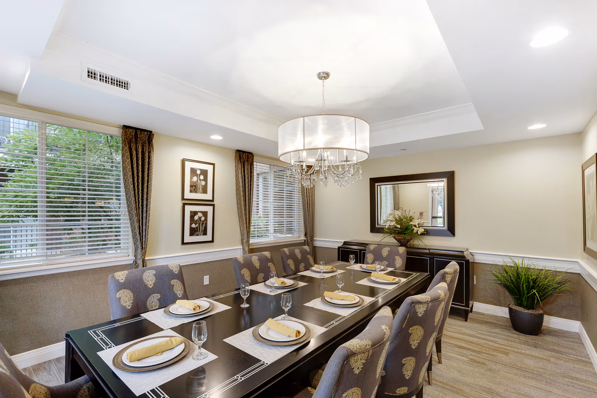 A well-lit dining room with a long dark wooden table set for eight people. Each place setting includes a plate, a napkin, and a glass. The chairs have patterned upholstery. There are two windows with blinds and curtains on the left wall, framed floral artwork between them, and a large mirror on the far wall above a sideboard with a floral arrangement. A modern chandelier hangs above the table, and there is a potted plant in the corner.