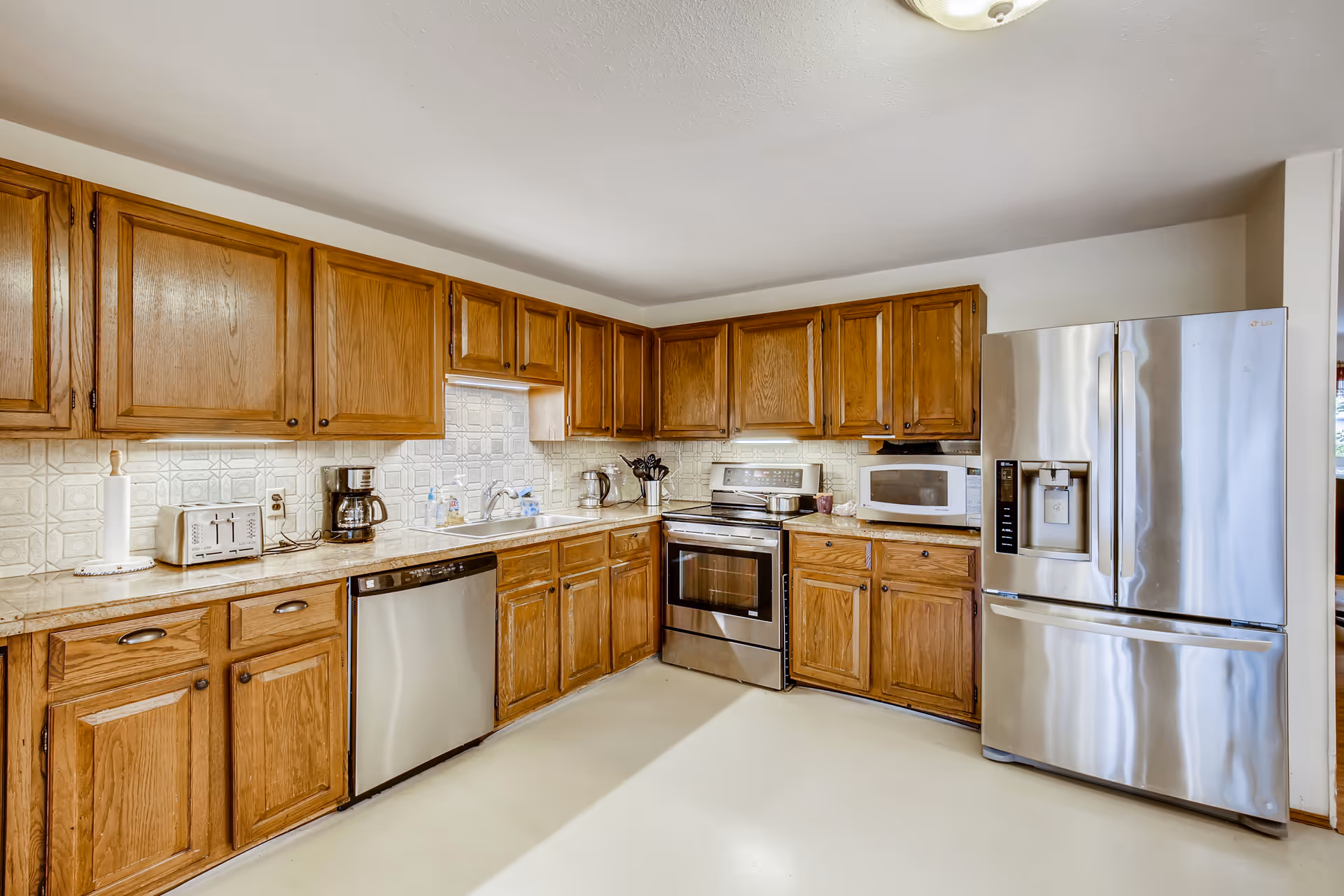 A spacious kitchen with wooden cabinets, a stainless steel refrigerator, dishwasher, oven, microwave, toaster, coffee maker, and a sink. The countertops are light-colored, and the backsplash has a geometric tile pattern. The floor is light-colored and the kitchen is well-lit.
