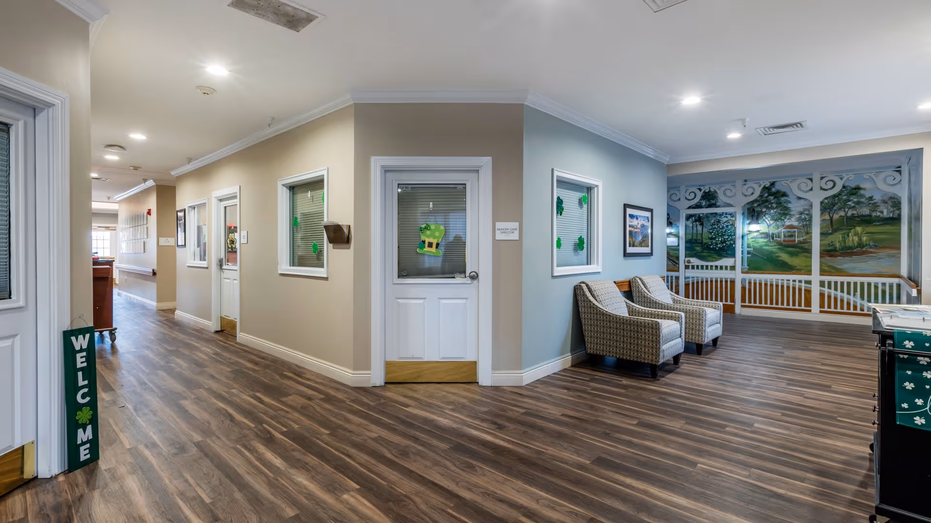 Interior view of a senior living facility hallway with wood flooring, beige and light blue walls, and white doors. Two patterned armchairs are placed near a wall with a large mural depicting a garden scene with trees and a gazebo. The doors and windows have St. Patrick's Day decorations with shamrocks and a leprechaun hat. A green welcome sign is visible near one door.