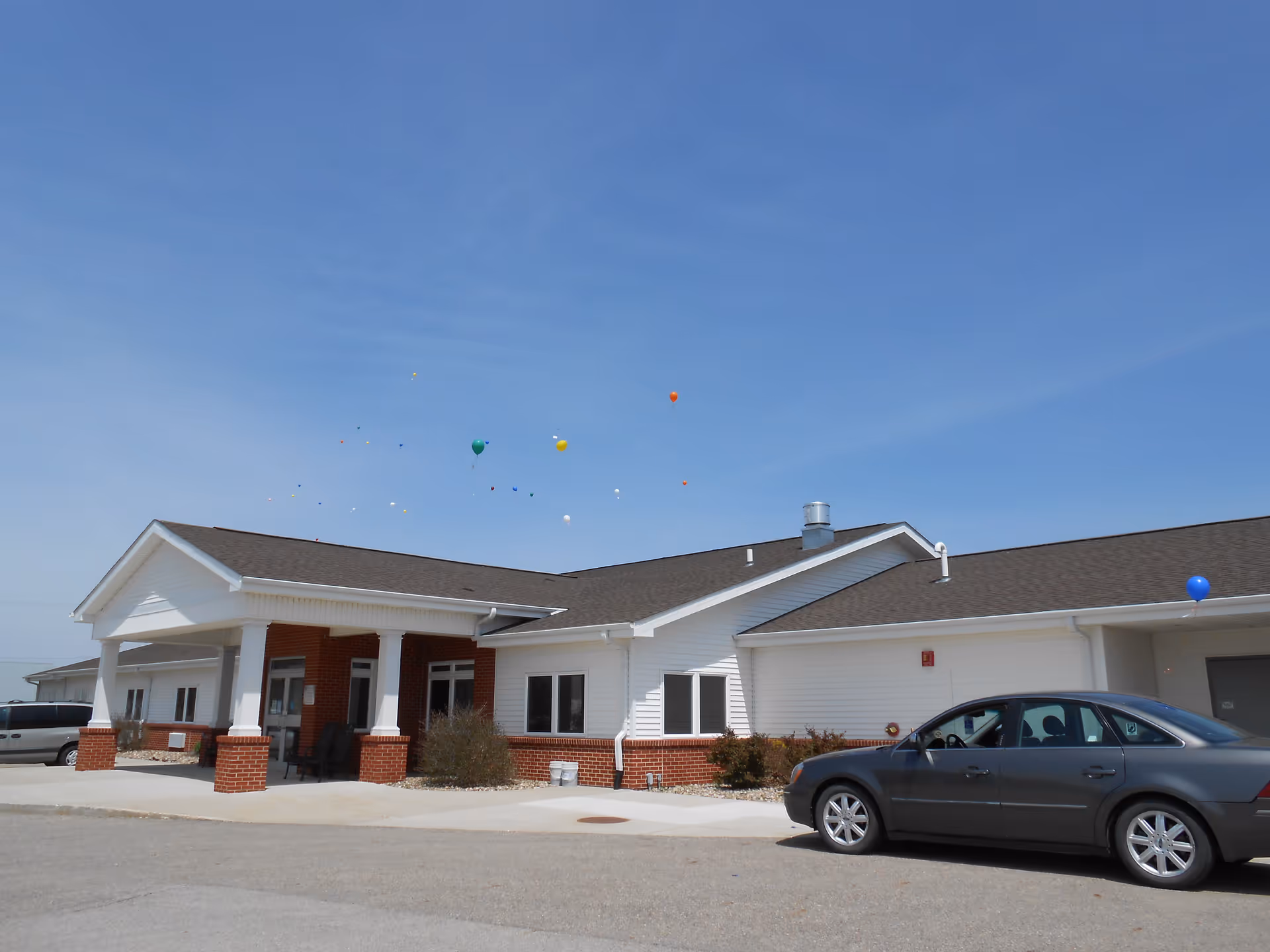 Exterior view of a single-story building with white siding and red brick accents under a clear blue sky. There is a covered entrance supported by white columns, a parked gray car in front, and several colorful balloons floating in the sky above the building.