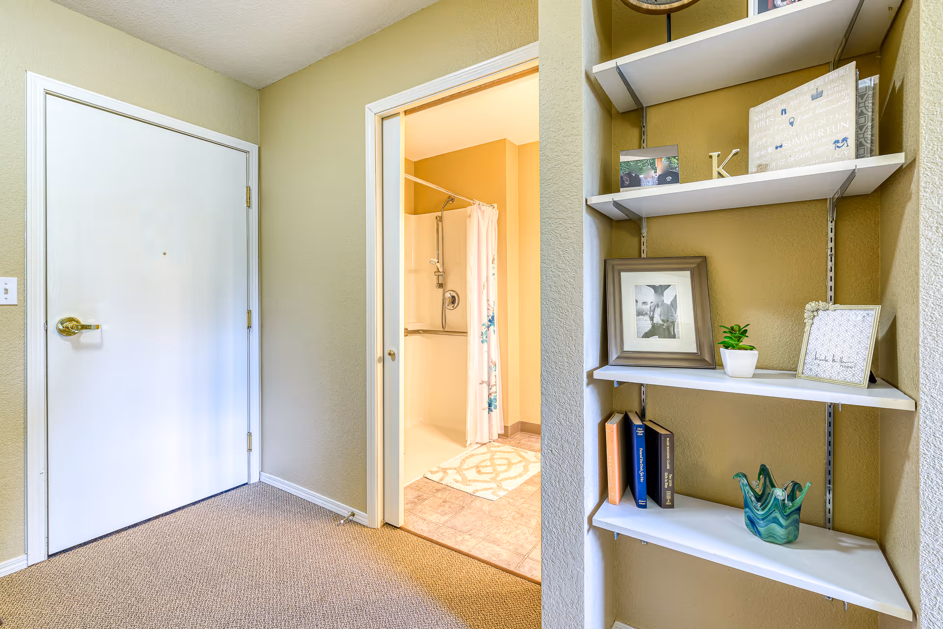 Interior view of a hallway leading to a bathroom with a shower and a floral shower curtain. To the right, there are white wall-mounted shelves holding framed photos, books, a small plant, and decorative items. The walls are painted beige and the floor is carpeted.