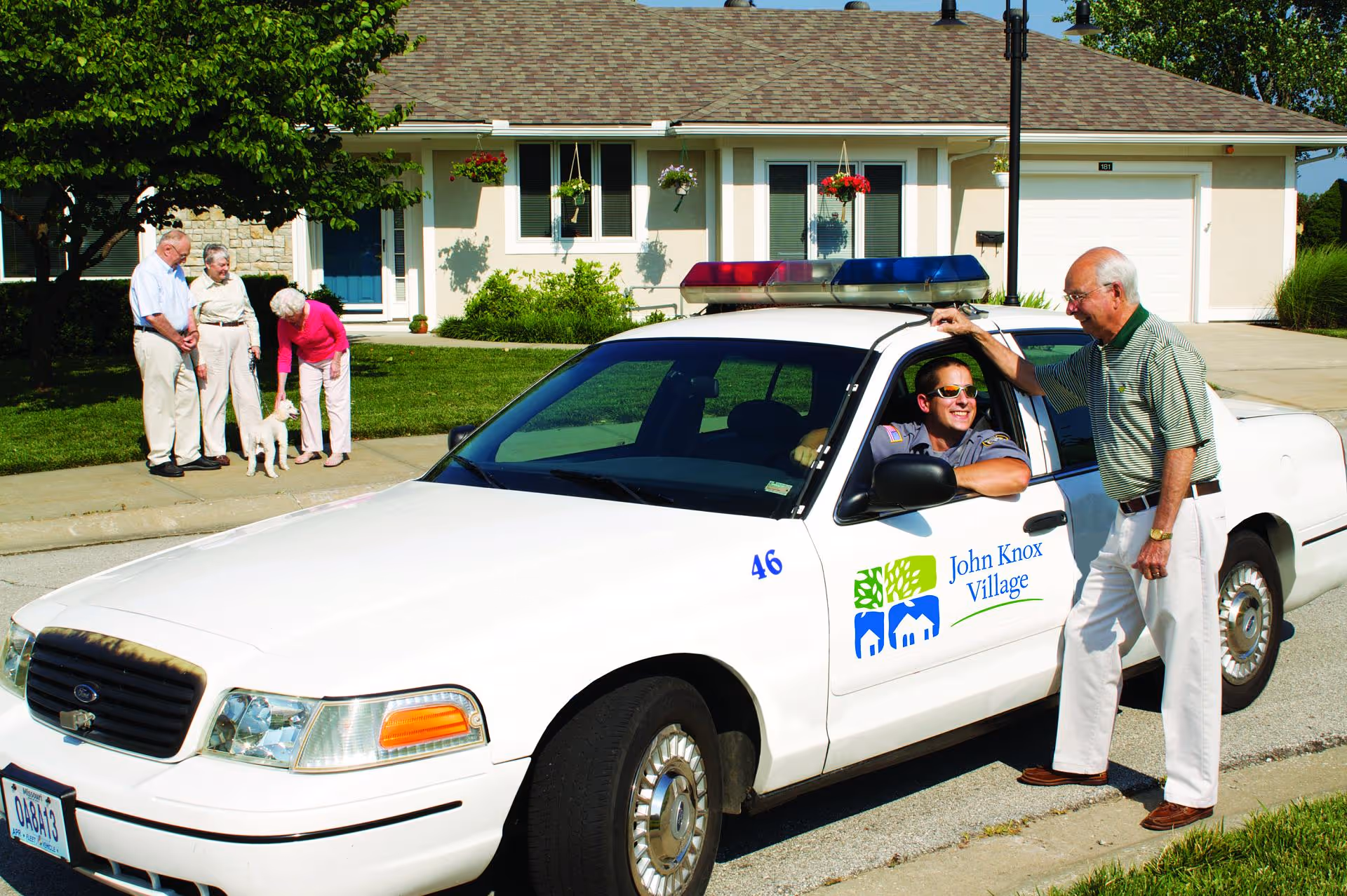 A John Knox Village patrol car parked in front of a single-story residence as a uniformed driver talks with an older man while other seniors and a small dog stand on the lawn.