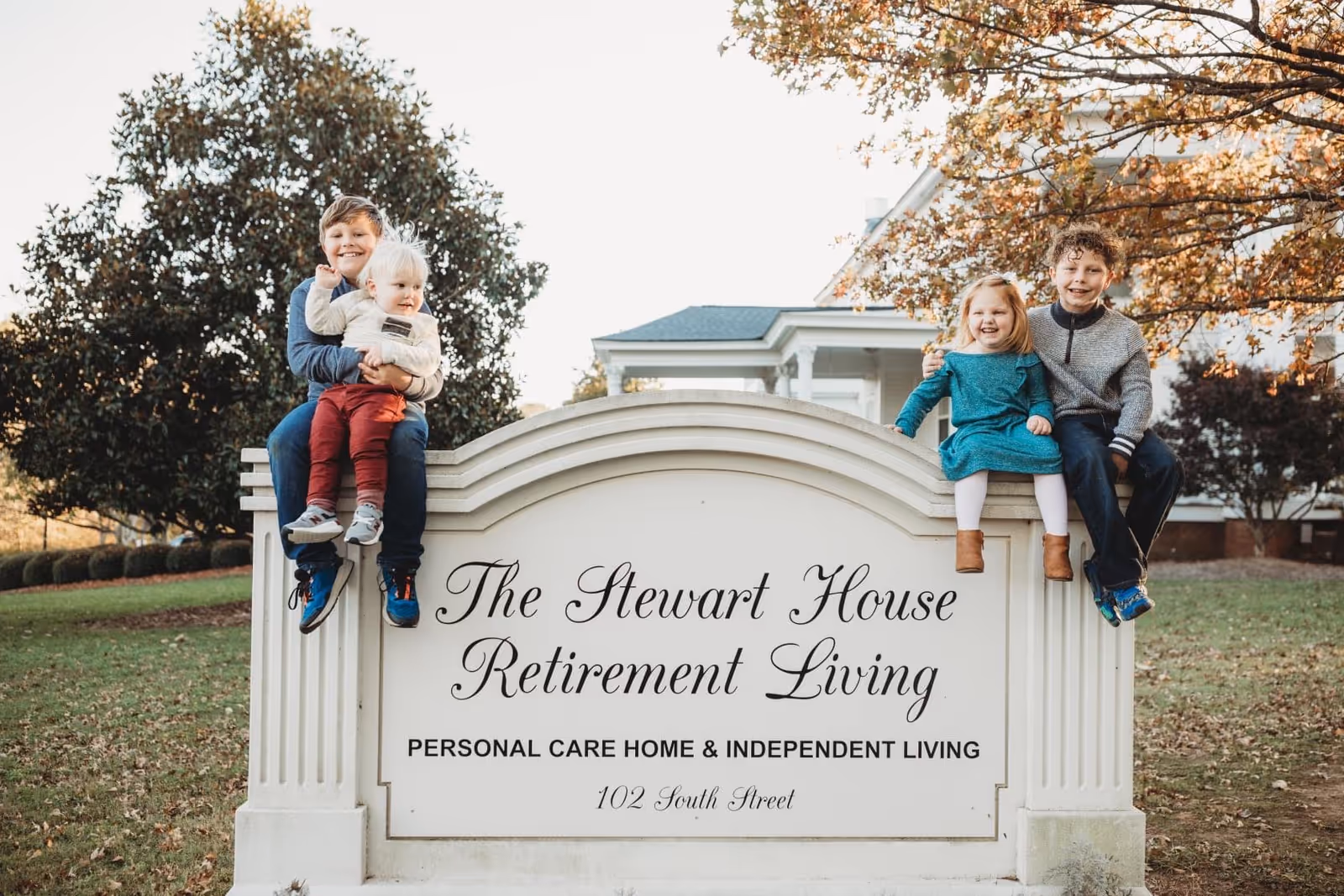 Children sit on a sign for The Stewart House Retirement Living in a sunny front yard.