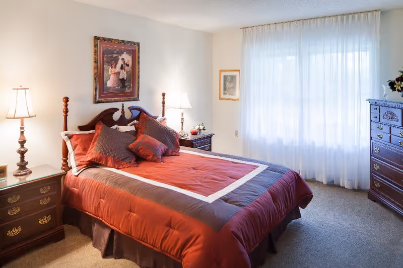 A well-lit bedroom with a large bed covered in a red and brown comforter and multiple pillows. There are two bedside tables with lamps on either side of the bed. A framed picture hangs above the headboard. A window with sheer white curtains allows natural light to fill the room. A wooden dresser with decorative items is positioned against the wall.