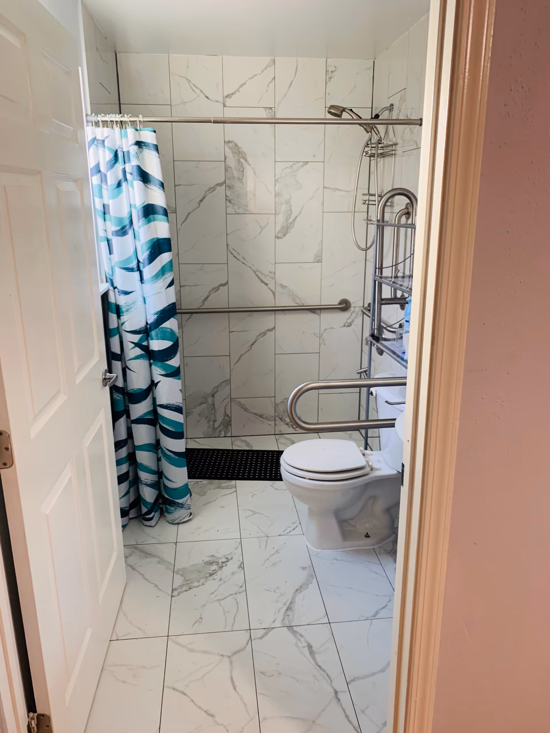 A bathroom with white marble-patterned tiles on the floor and walls. There is a white toilet with a safety grab bar on the right side. A shower area with a blue and white patterned shower curtain is visible on the left side. The shower has a metal grab bar and a handheld showerhead. A metal shelving unit is placed next to the toilet.