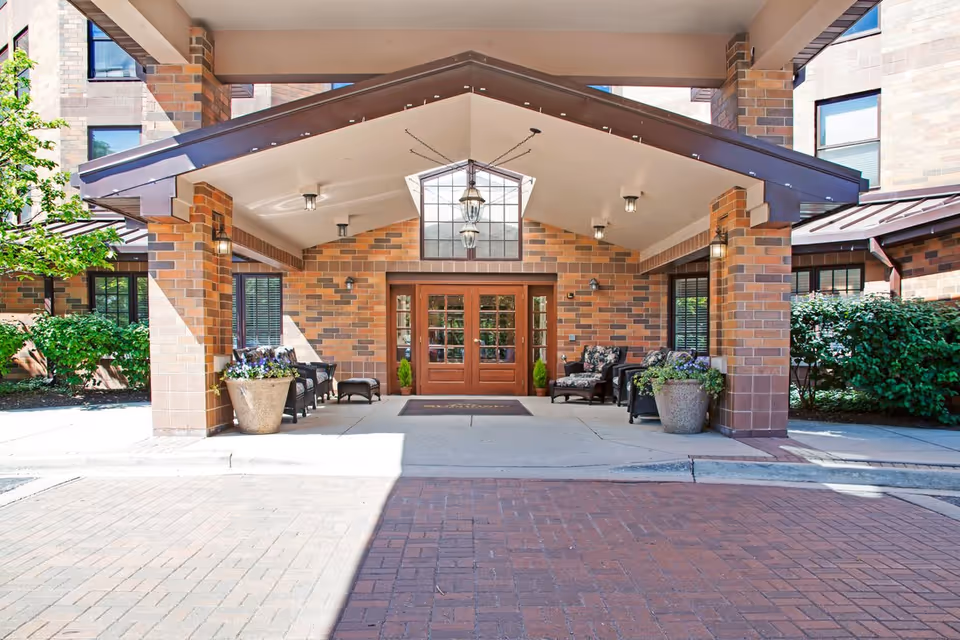 Entrance of a senior living facility with a covered porch supported by brick columns. There are wooden double doors with glass panels, outdoor seating with cushioned chairs, potted plants, and hanging lantern-style lights under the porch roof.