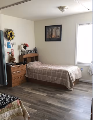 A simple bedroom with a single bed covered in a beige plaid blanket. Next to the bed is a wooden dresser with a small Christmas tree decoration, a lamp, and framed photos. A framed picture hangs on the wall above the bed. There is a window with white curtains letting in natural light. The floor is wood laminate.