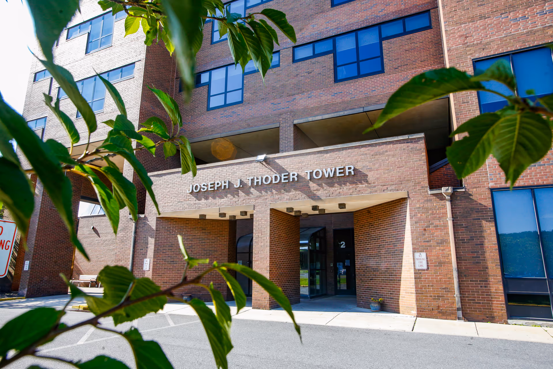 Exterior view of the Joseph J. Thoder Tower building at Gracedale Nursing Home, framed by green leaves in the foreground. The building is made of red brick with large windows and an entrance under a covered area.