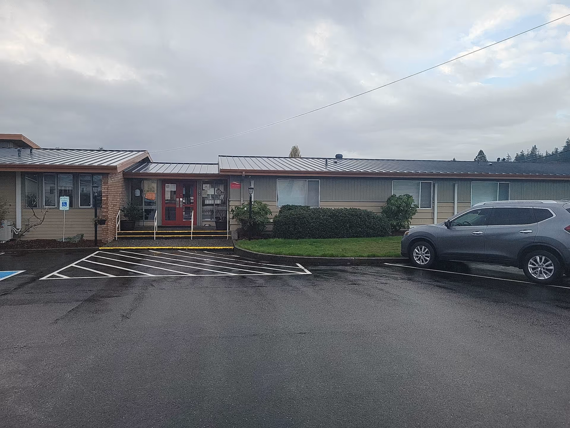 Exterior view of a single-story building with a metal roof and beige siding, featuring a red double-door entrance with steps and a ramp. There is a parking lot in front with a gray SUV parked on the right and a handicapped parking sign on the left. The sky is cloudy.