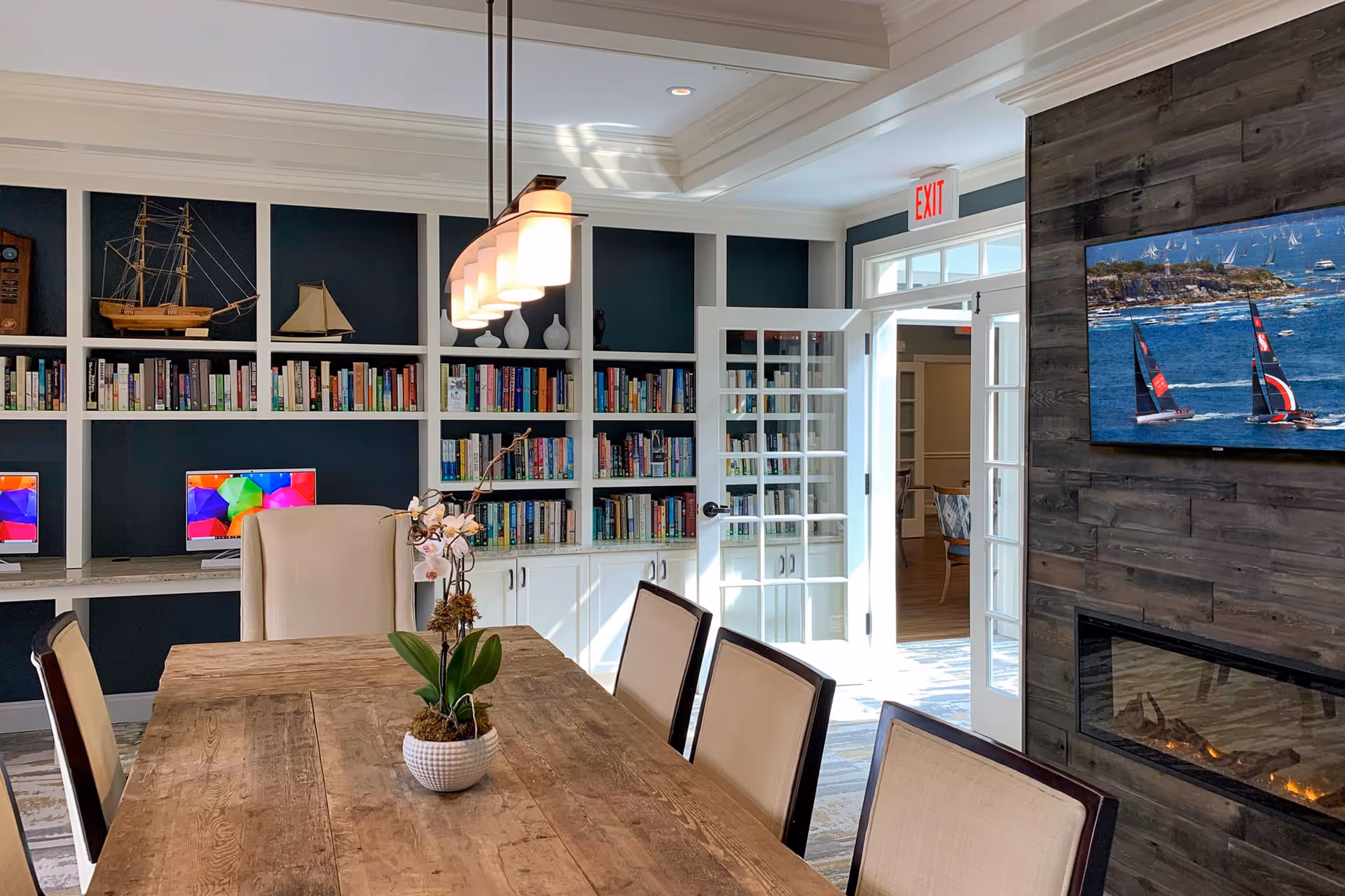 A bright and cozy common area with a wooden table surrounded by chairs, a modern hanging light fixture above, built-in bookshelves filled with books and decorative items, two computer monitors on a desk, a glass door leading to another room, and a wall-mounted TV above a fireplace displaying a sailing scene.