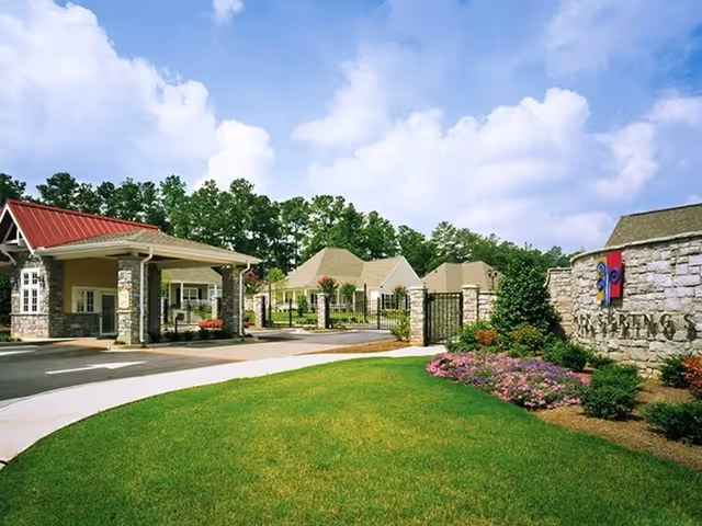 Entrance to a senior living facility with a gated driveway, a guardhouse with a red roof, stone pillars, and well-maintained landscaping including green grass and colorful flowers under a partly cloudy blue sky.