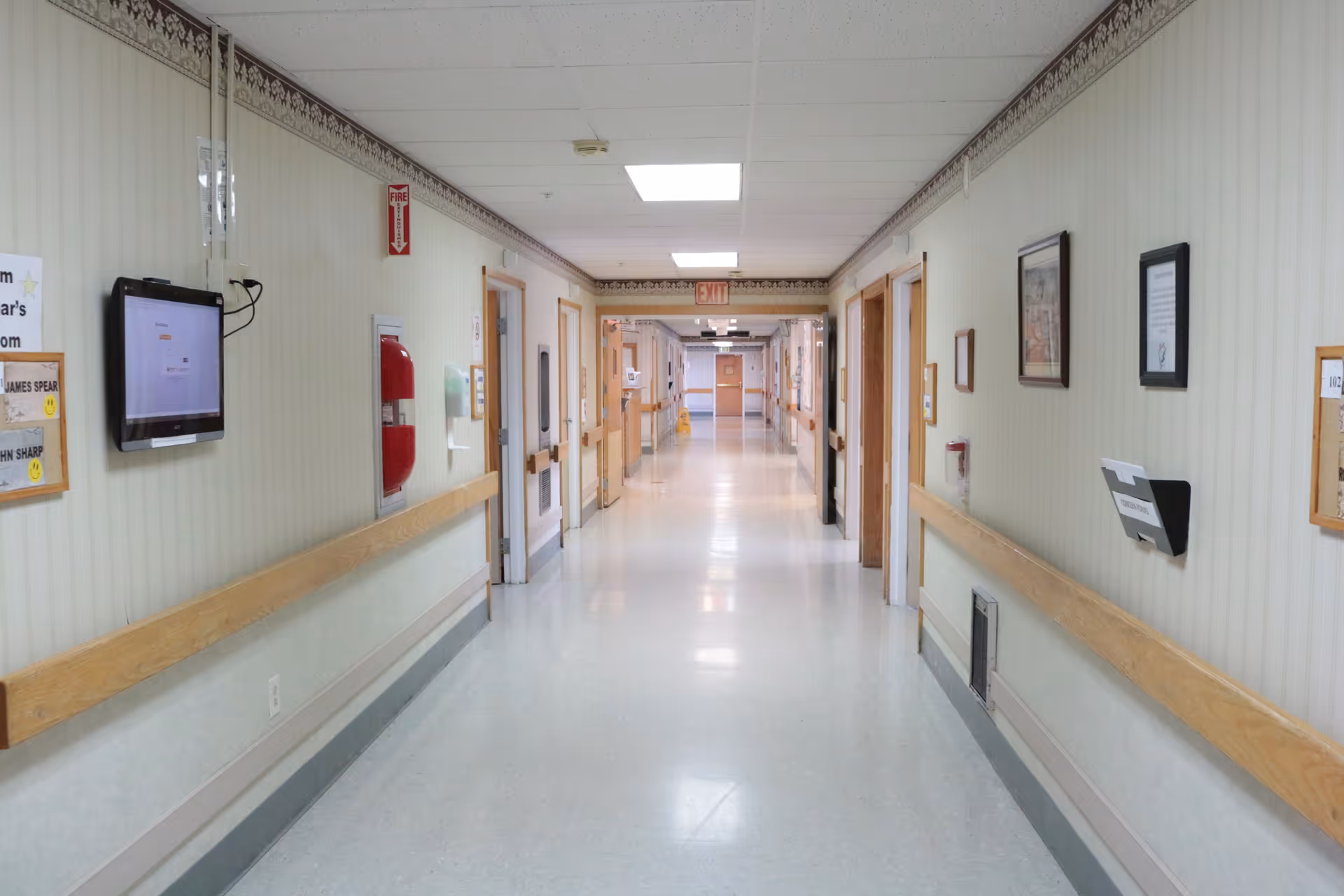 A long, clean hallway in a senior living facility with light-colored walls, wooden handrails on both sides, several closed doors, framed pictures and notices on the walls, a fire extinguisher, and ceiling lights. The hallway extends into the distance with an exit sign visible at the far end.