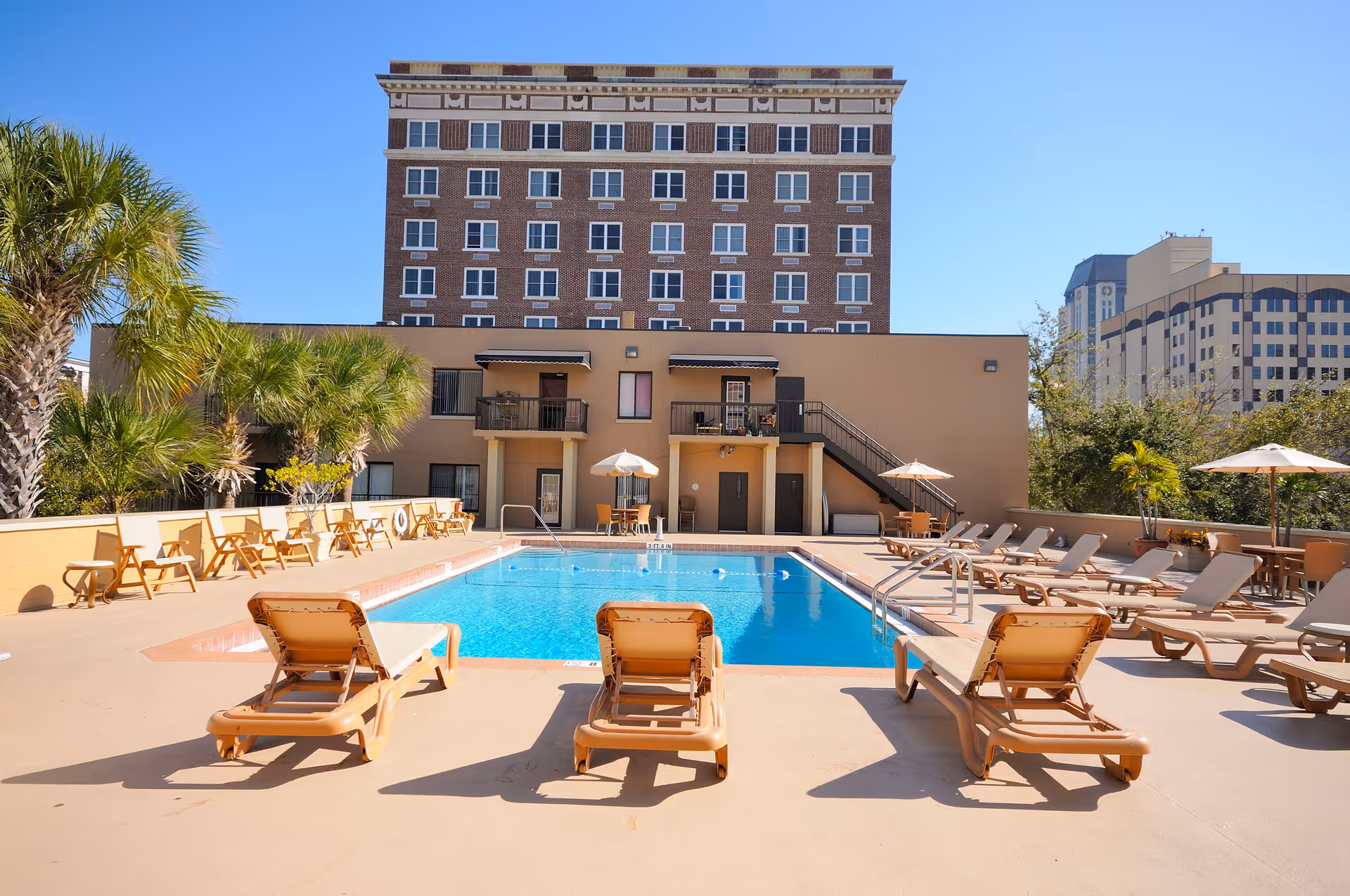 Outdoor swimming pool area with several beige lounge chairs and umbrellas around it. Palm trees and other greenery are visible on the left and right sides. A multi-story brick building and other buildings are in the background under a clear blue sky.