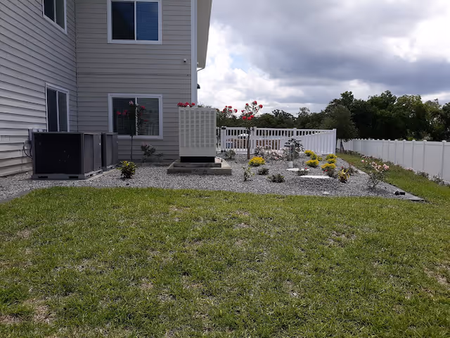 Outdoor area beside a building showing a small garden with various plants and flowers, a patch of green grass in the foreground, and several air conditioning units placed on gravel near the building. A white fence runs along the right side and background of the garden area under a partly cloudy sky.