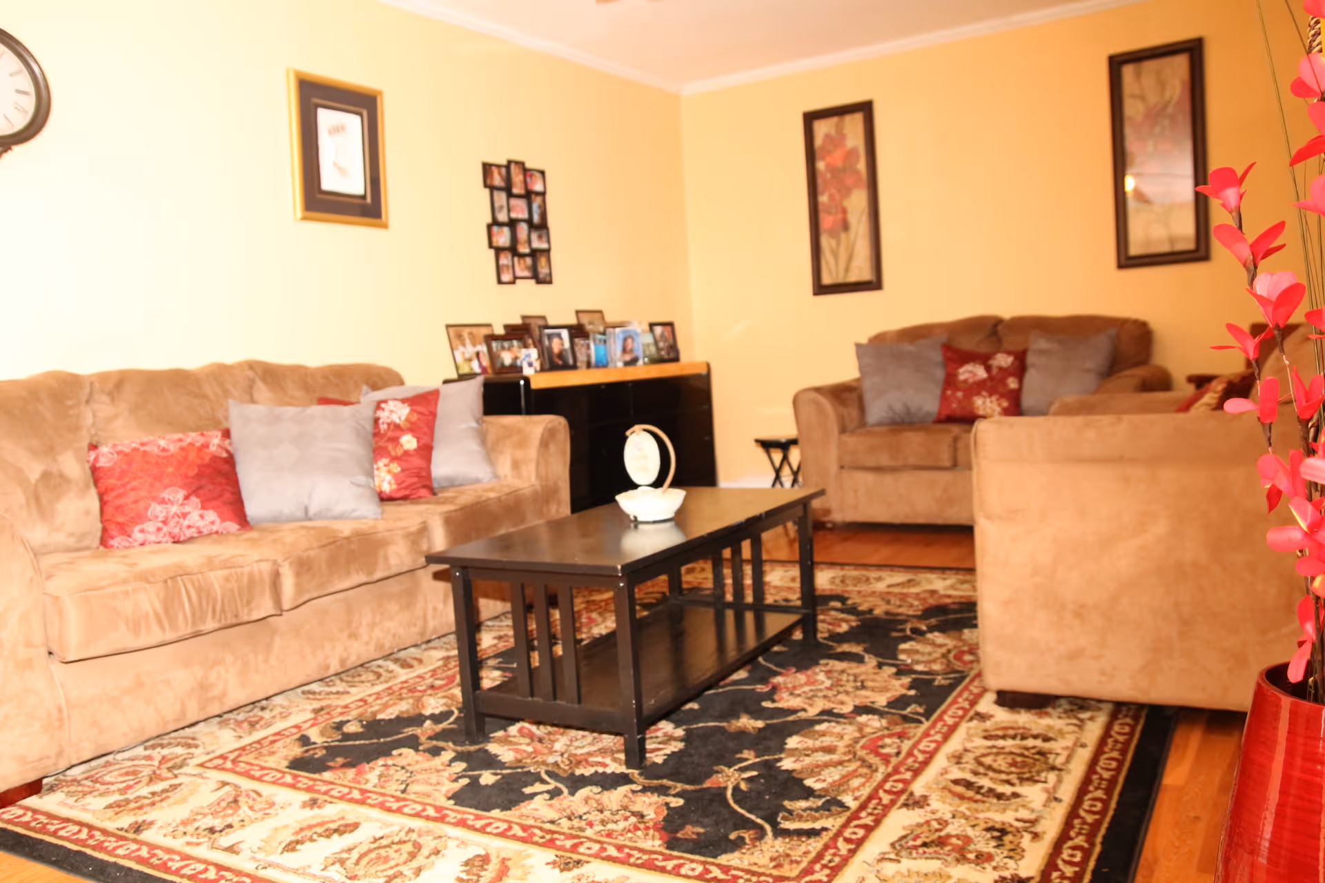 A cozy living room with beige walls and a patterned area rug. There are two brown sofas and one brown armchair, each adorned with decorative pillows in red and gray. A black coffee table sits in the center with a small decorative item on it. The walls feature framed artwork and a clock, and a black cabinet holds several framed photos. A red vase with pink flowers is visible on the right side of the image.
