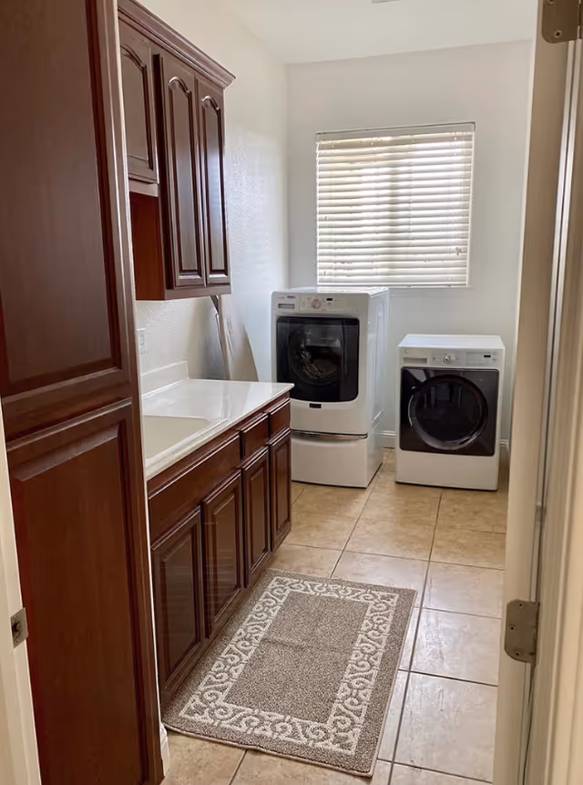 Laundry room with front-loading washer and dryer, wooden cabinets and countertop with sink, tiled floor, and a window with blinds.