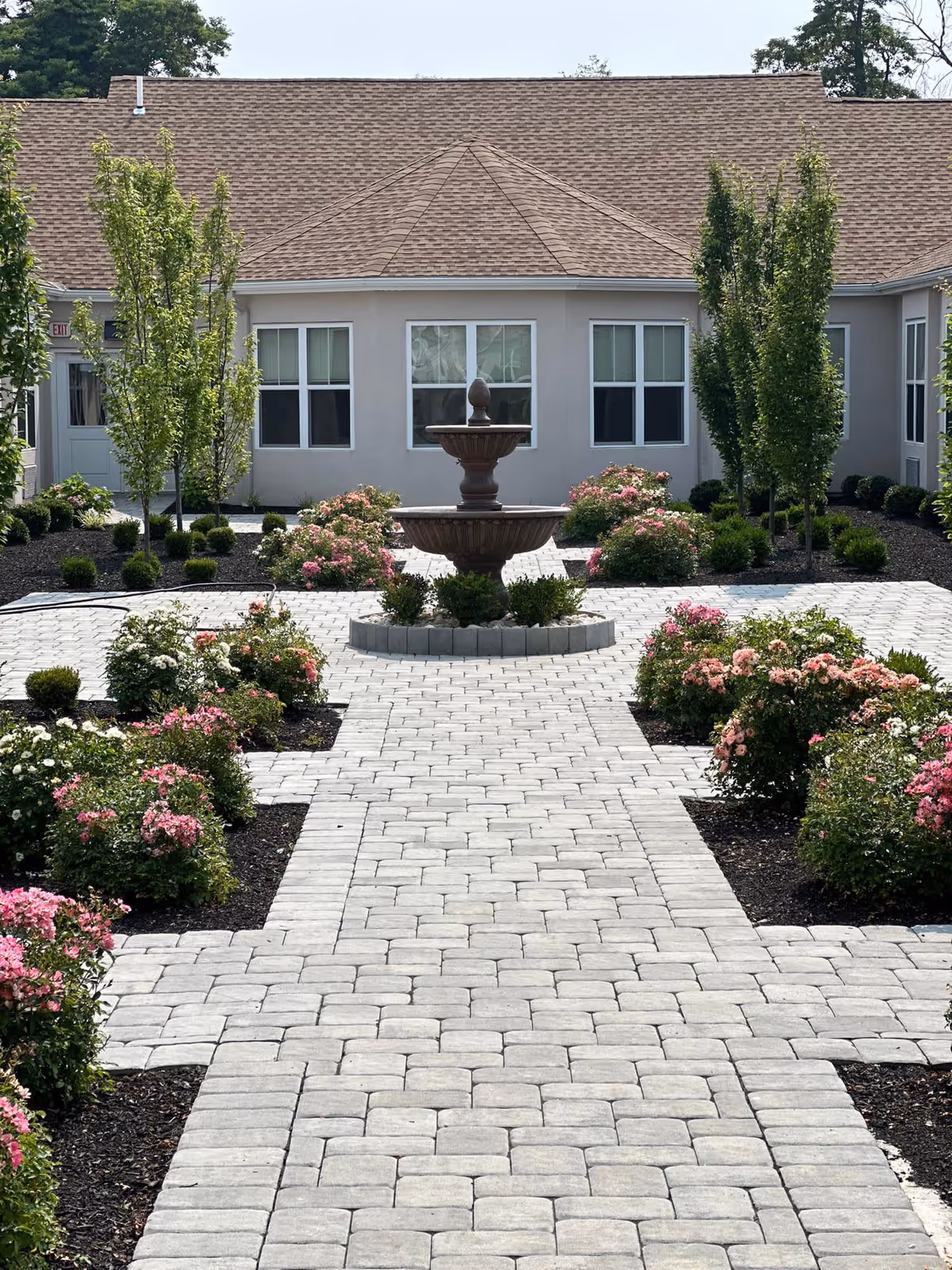 A landscaped outdoor courtyard at Woolwich Gardens Assisted Living featuring a central two-tiered stone fountain surrounded by neatly arranged flower beds with pink and white flowers, small bushes, and young trees. The courtyard has a paved stone walkway leading to a building with multiple windows and a brown shingled roof.