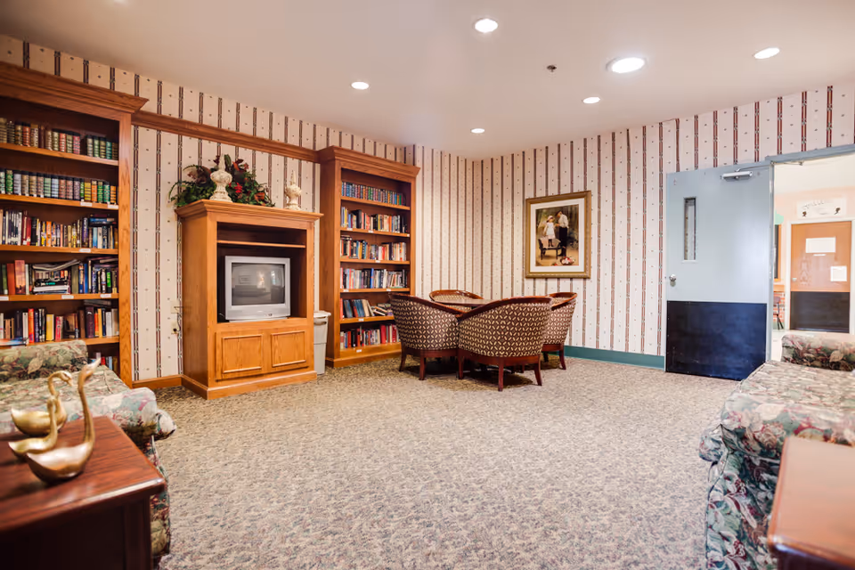 A cozy living room area in a senior living facility featuring floral patterned sofas, a wooden entertainment center with a TV, two tall bookshelves filled with books, a round table with four upholstered chairs, patterned wallpaper, and a framed picture on the wall. The room has carpeted flooring and a partially open door leading to another area.