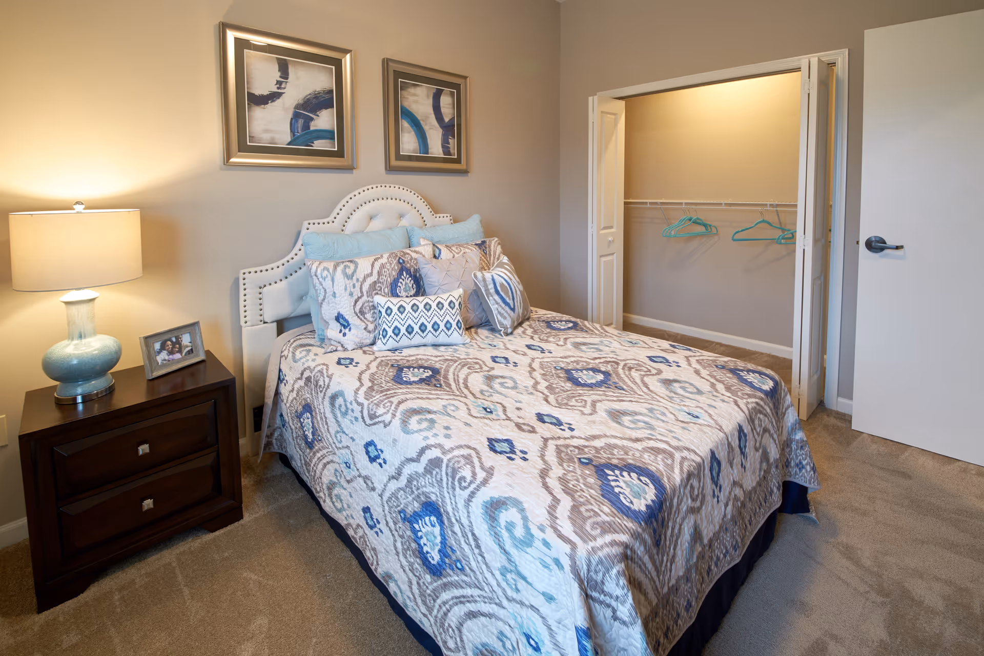 A neatly made bed with a patterned blue, gray, and white comforter and multiple pillows in a bedroom. There is a wooden nightstand with a blue lamp and a framed photo next to the bed. Two abstract paintings hang on the beige wall above the bed. An open closet with a few hangers is visible, along with a partially open white door.