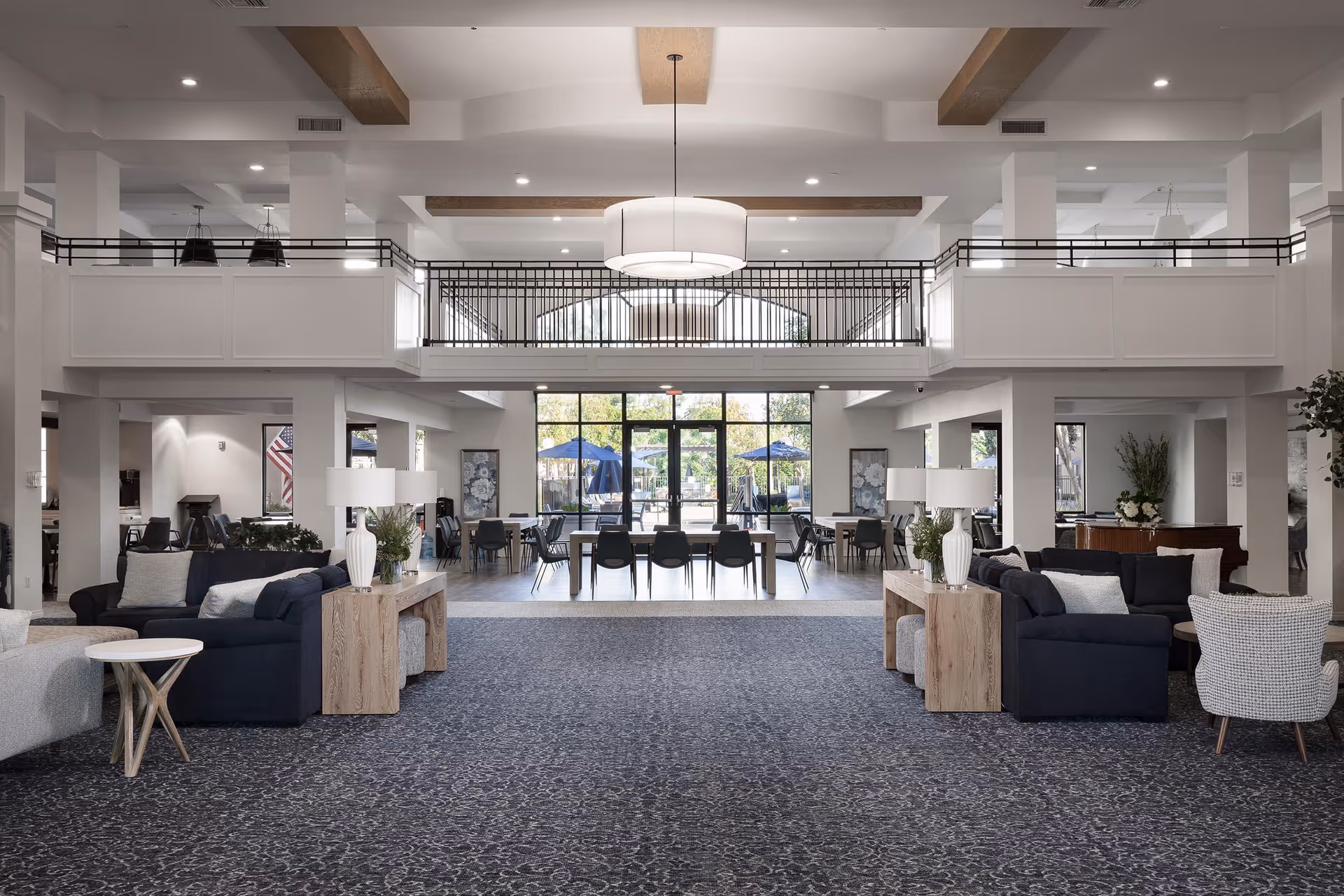 Spacious and modern senior living facility common area with high ceilings, a large hanging light fixture, multiple seating arrangements including sofas and chairs, wooden side tables with lamps and plants, and large windows and glass doors leading to an outdoor patio with umbrellas.