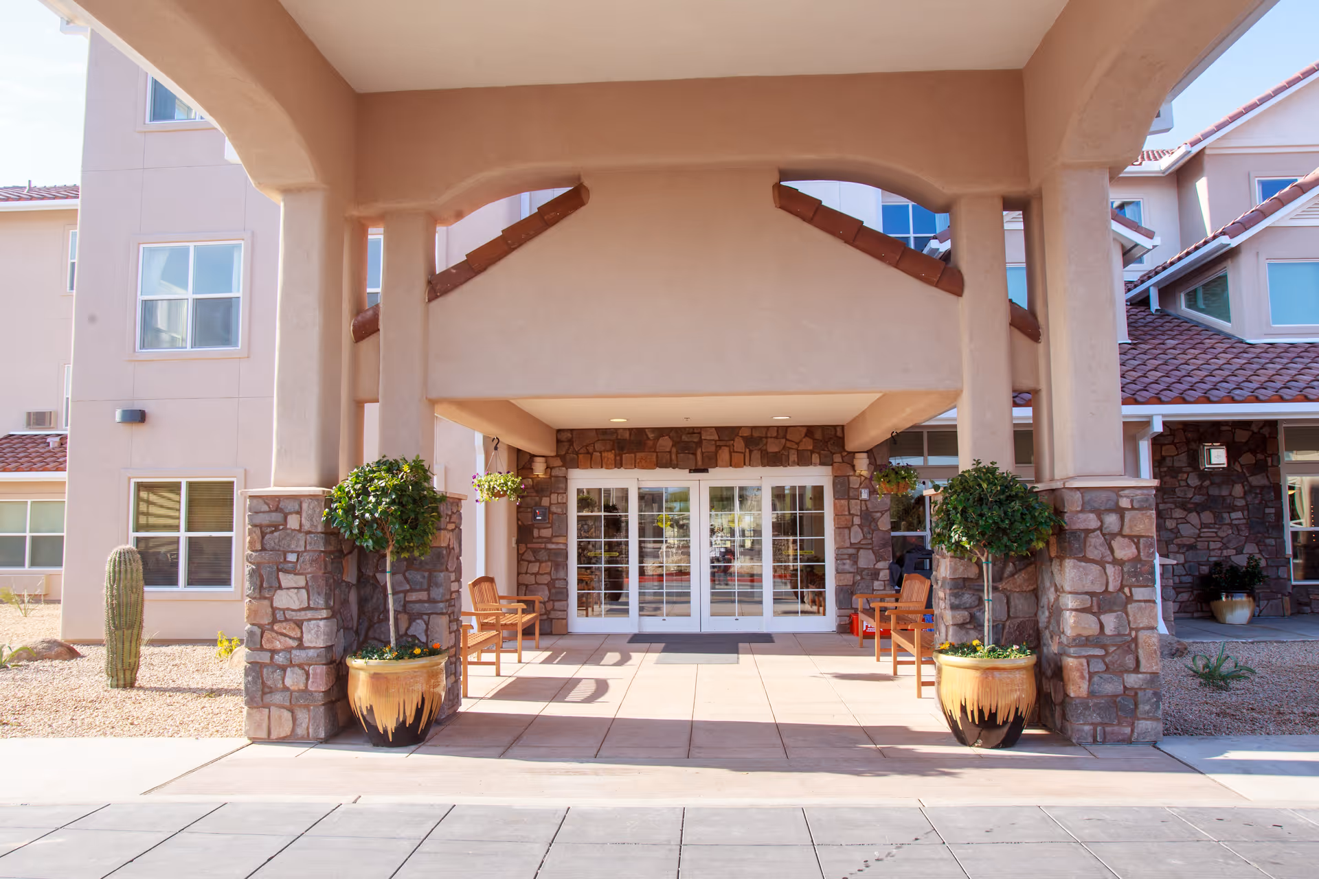 Entrance of a senior living facility with a covered porch supported by stone pillars, potted plants, wooden benches, and glass double doors leading inside. The building has a beige exterior with red tile roofing.