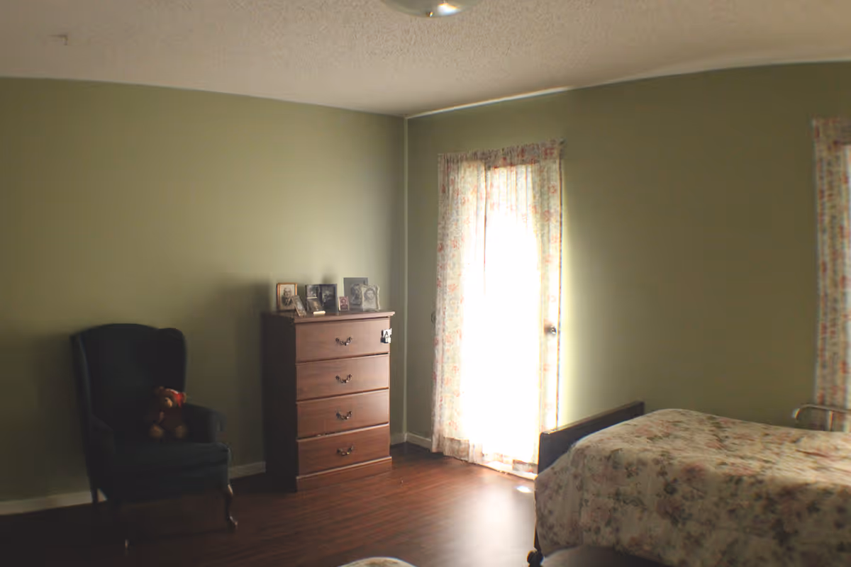 A bedroom with light green walls, a wooden dresser with framed photos on top, a dark upholstered armchair with a teddy bear sitting on it, and a bed with floral bedding. Two windows with floral curtains allow natural light into the room.