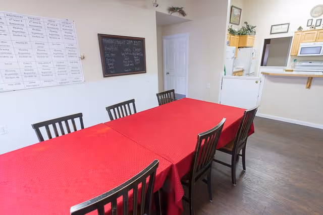 A dining area with a long table covered by a red tablecloth and six wooden chairs around it. On the wall, there is a weekly activity schedule and a blackboard with daily menu items written on it. In the background, there is a kitchen area with wooden cabinets, a white refrigerator, microwave, and a counter with a pass-through window.